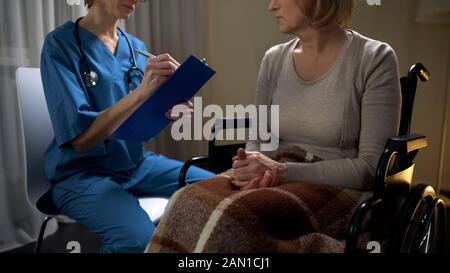 Doctor on call talking to sick woman in wheelchair and making notes, diagnosis Stock Photo