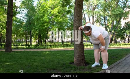 Fat man tired after running in park, leaning on tree, tiresome workouts outdoors Stock Photo