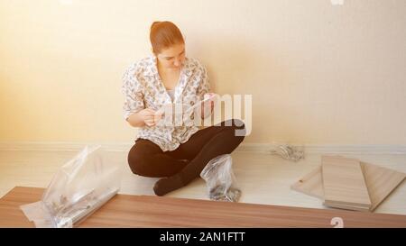 serious housewife sits on floor and reads instruction looking at cabinet furniture fasteners in apartment kitchen sunlight Stock Photo