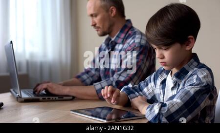 Kid scrolling on tablet, father working on laptop computer, lack of attention Stock Photo