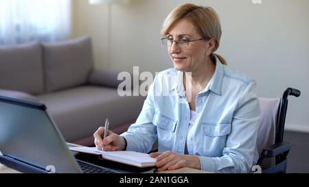Senior woman in wheelchair studying online and making notes, video lessons Stock Photo