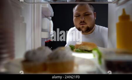 Overweight young man opening fridge at night to take big burger, calories Stock Photo