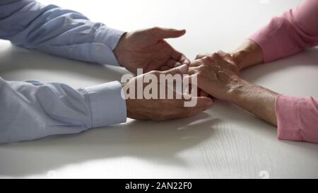 Old male taking his wifes hands, couple sitting at table together, love and care Stock Photo