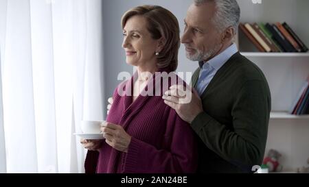 Senior couple looking in window, male hugging lady shoulders from behind, family Stock Photo