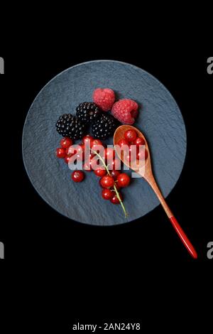 Red currants, blackberries and raspberries on black plate with wooden spoon, Germany Stock Photo