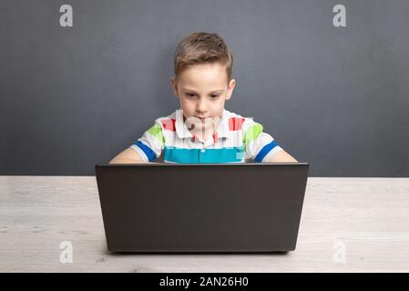 Boy works on a laptop computer and a desk. Concept of using computers for gaming and learning. Front view. Clean wall with space for text in backgroun Stock Photo