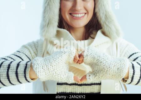Closeup on happy trendy female in white striped sweater, scarf and ear flaps hat showing heart shaped hands on winter light blue background. Stock Photo