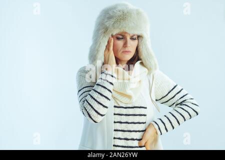 stressed modern woman in white striped sweater, scarf and ear flaps hat having headache isolated on winter light blue background. Stock Photo