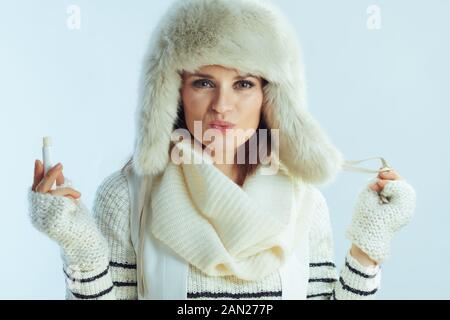Portrait of modern woman in white striped sweater, scarf and ear flaps hat using hygienic lipstick isolated on winter light blue background. Stock Photo