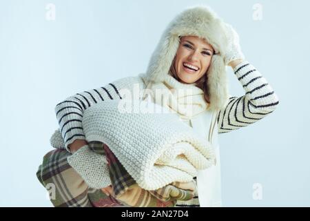 Portrait of smiling middle age woman in white striped sweater, scarf and ear flaps hat holding a stack of warm blankets on winter light blue backgroun Stock Photo