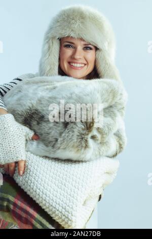 Portrait of smiling elegant female in white striped sweater, scarf and ear flaps hat holding a stack of warm blankets isolated on winter light blue ba Stock Photo