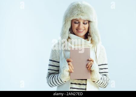 smiling young woman in white striped sweater, scarf and ear flaps hat looking at a book isolated on winter light blue background. Stock Photo