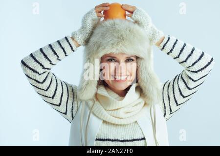 Portrait of happy modern female in white striped sweater, scarf and ear flaps hat holding orange on head against winter light blue background. Stock Photo