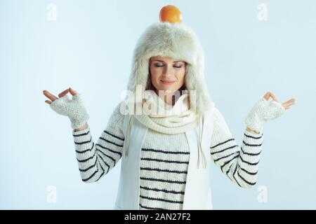 relaxed elegant female in white striped sweater, scarf and ear flaps hat with orange on head meditating against winter light blue background. Stock Photo
