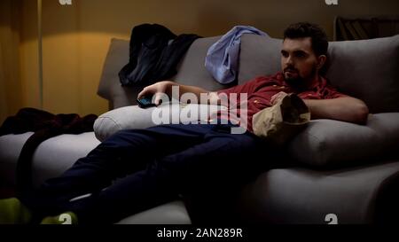 Messy man lying on sofa, watching tv, covered with chips crisps, untidiness Stock Photo