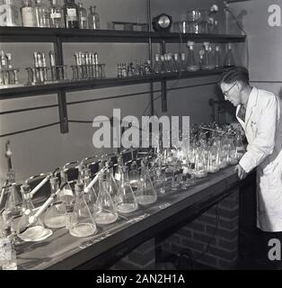 1950s, historical, male research scientist in a white-coat studying a collection of glass test tubes in a laboratory, Leeds University, with the words 'Leave Taps On'' written on the workbench. Stock Photo