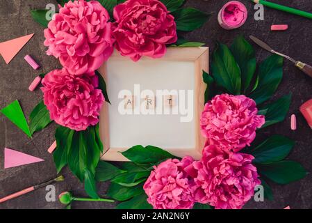 Compostion of ART word typing on wooden blocks on white card in photo frame, pink peony flowers with green leaves surrounded by variety of drawing sup Stock Photo
