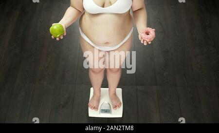 Overweight girl standing on scales with apple and donut, food choice dieting Stock Photo