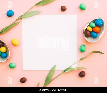Chocolate Easter Eggs with colorful candies over pink background, flowers and copy space for text, white blank mockup sheet of paper Stock Photo