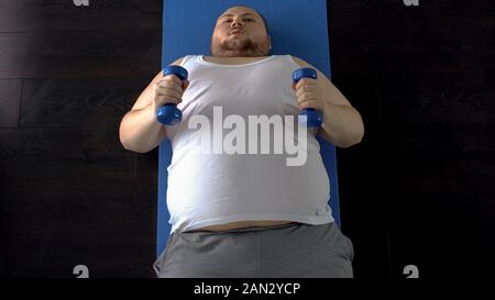 Purposeful fat man lifting dumbbells with great effort lying on mat, top view Stock Photo