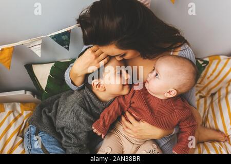 Young beautiful mother with her children on the bed, tightly hugs her son and little daughter, kisses her forehead, children laugh Stock Photo