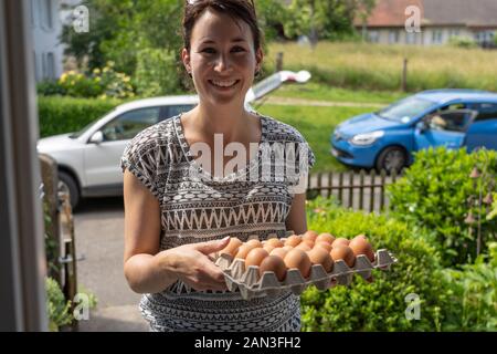 Beautiful young natural woman friendly smiling, delivers fresh brown eggs from free range chicken in egg box. Her car stands with open trunk in the st Stock Photo