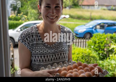 Beautiful young natural woman friendly smiling, delivers fresh brown eggs from free range chicken in egg box. Her car stands with open trunk in the st Stock Photo