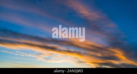 Panoramic orange illuminated sky background in evening dusk view. Dramatic sunset on blue striped cloudscape with sunlit fiery clouds. Eco and weather. Stock Photo