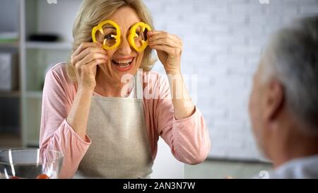 Playful woman making faces with pepper slices, senior couple having fun at home Stock Photo