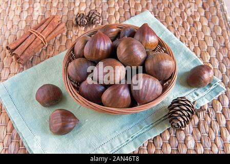 Sweet Chestnuts (Castanea sativa) in small wicker basket with pine cones on vintage green napkin Stock Photo