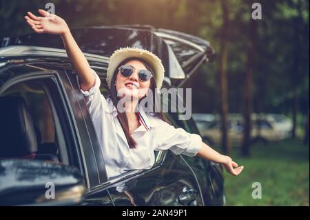 Happy woman waving hand outside open window car with meadow and mountain forest background. People lifestyle relaxing as traveler on road trip in holi Stock Photo