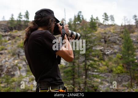 A low angle and rear view of an animal and nature photographer taking photos in rural landscape, with blurry cliff and pine trees in background Stock Photo