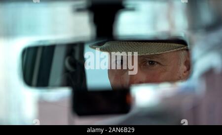 Elderly man wearing hat and looking in rear view car mirror, driver close-up Stock Photo