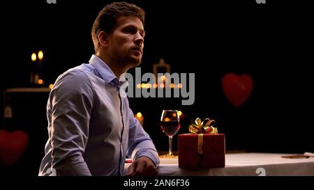 Upset man sitting in restaurant with red gift box, girlfriend missing date Stock Photo