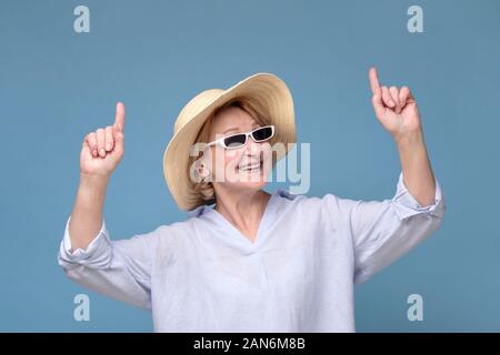 Senior woman in summer hat and sunglasses pointing up having great mood smiling studio isolated on blue wall. Ready for travel on summer vacation Stock Photo