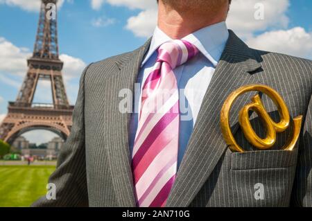 Unrecognizable businessman standing in front of the Eiffel Tower with a golden at symbol sticking out of the lapel pocket of his pinstripe suit in Par Stock Photo