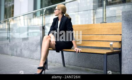 Happy businesswoman sits on bench, pleased with successful working day, rest Stock Photo