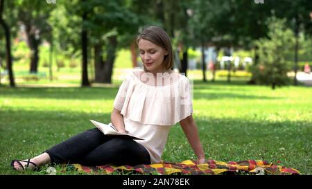 Pretty lady reading romantic novel, sitting on plaid in park, book lover Stock Photo