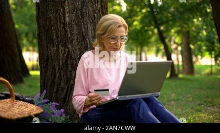 Beautiful mature woman inserting card number on laptop pc in park, shopping Stock Photo
