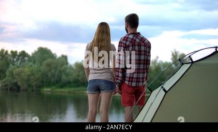 Young couple looking at river in campsite, standing near tent, back view Stock Photo