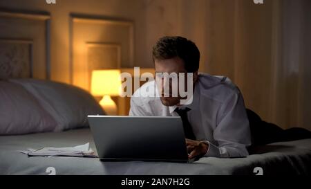 Serious man lying on bed and improving business knowledge online using laptop Stock Photo