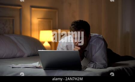 Exhausted engineer lying on bed, working on laptop at night, reading mail Stock Photo