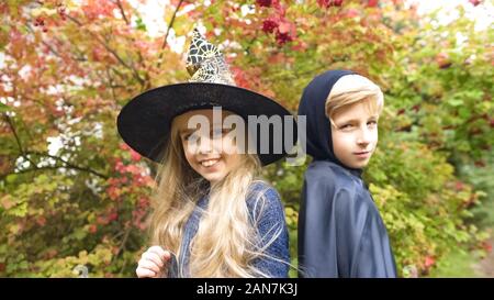 Cute girl in witch hat and boy in mantle posing for camera in forest, Halloween Stock Photo