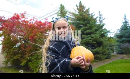 Happy girl holding pumpkin and posing for camera in Halloween cat costume Stock Photo