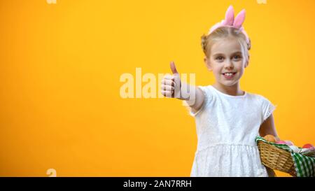 Female child in bunny ears headband holding Easter basket and showing thumbs up Stock Photo
