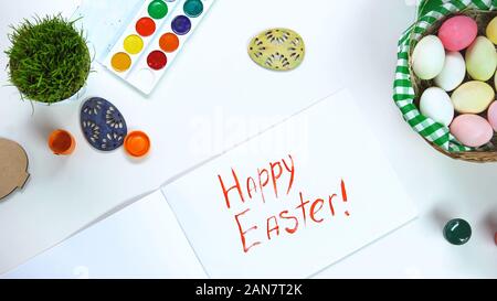 Happy Easter phrase written on paper, table with decorative eggs and paints Stock Photo