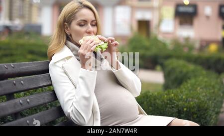Hungry pregnant woman enjoying delicious burger sitting on bench in park Stock Photo