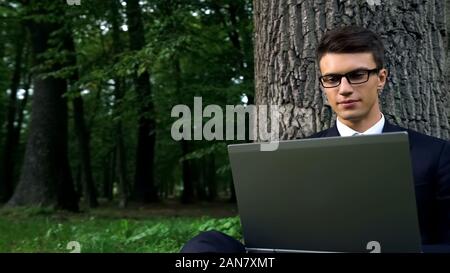 Serious business man working on laptop, sitting under tree in central park Stock Photo