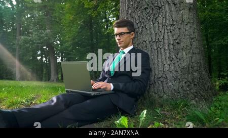 Young businessman with laptop sitting under tree, working remotely in park Stock Photo