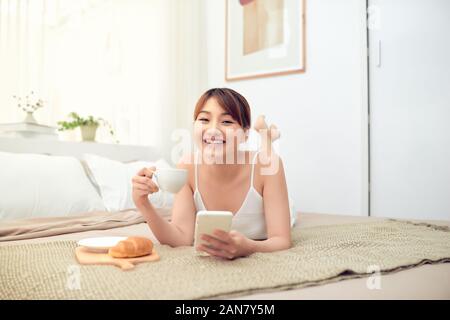 Cheerful young Asian woman using mobile phone lying on bed in morning Stock Photo